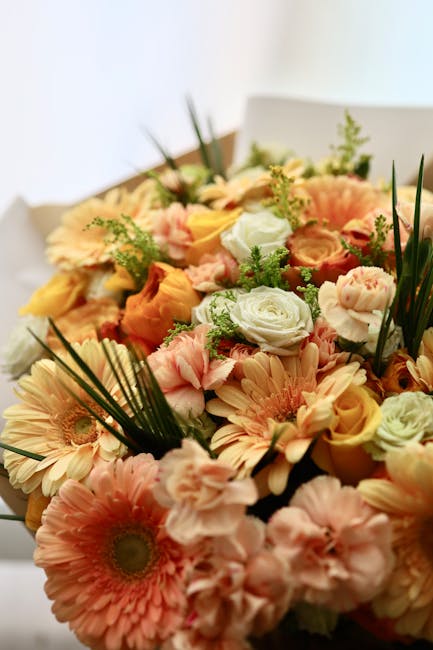 A close-up of a floral bouquet featuring a variety of fresh flowers including peach and cream roses, pink gerbera daisies, and light pink ranunculus, accented with green foliage and long, slender palm