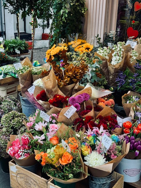 A vibrant display of freshly prepared flower bouquets arranged on outdoor tables in front of a floral shop. The bouquets feature a variety of flowers, including bright yellow sunflowers, pink tulips, 