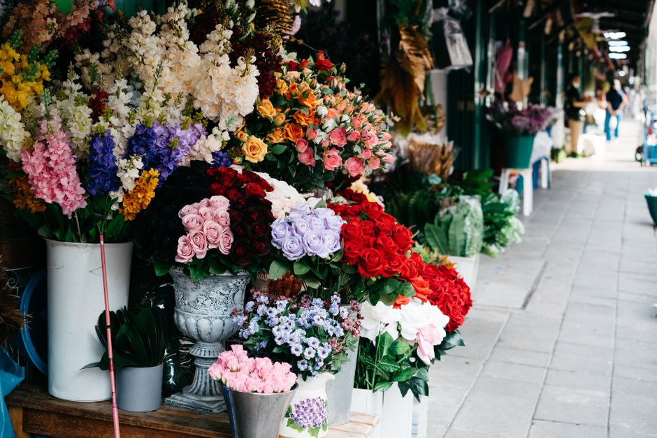 A vibrant outdoor display of fresh floral arrangements and bouquets on a flower shop's street stall. The flowers include a variety of roses in pink, red, purple, and peach tones, alongside white lilie